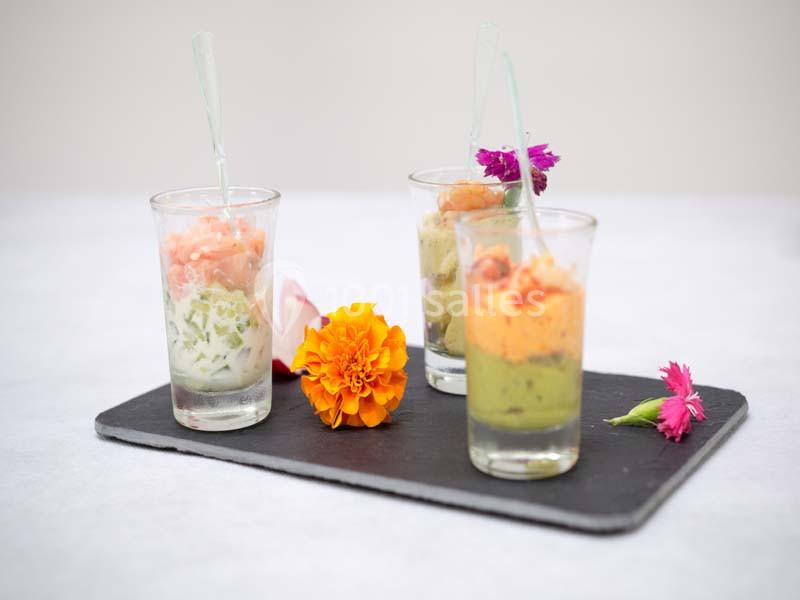 Trois verrines colorées garnies de préparations variées, présentées sur une ardoise avec des fleurs décoratives.