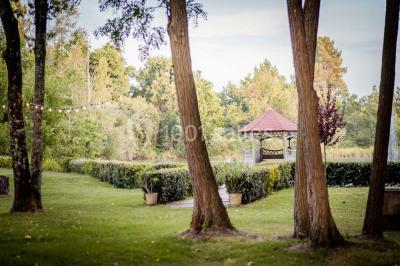 Un kiosque blanc au bord d'un étang avec une fontaine, entouré d'arbres et d'une réception en arrière-plan.
