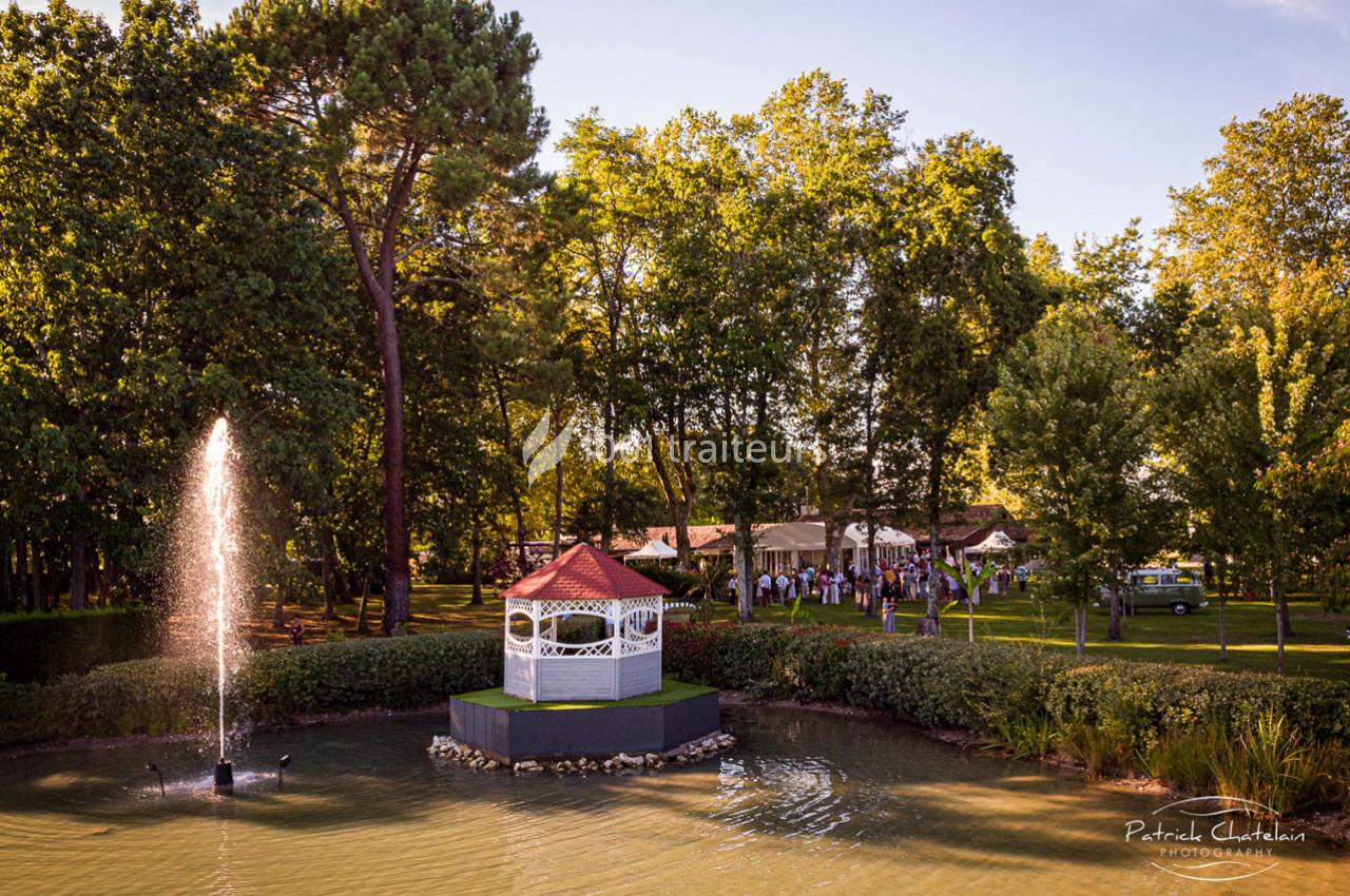 Un kiosque blanc au bord d'un étang avec une fontaine, entouré d'arbres et d'une réception en arrière-plan.