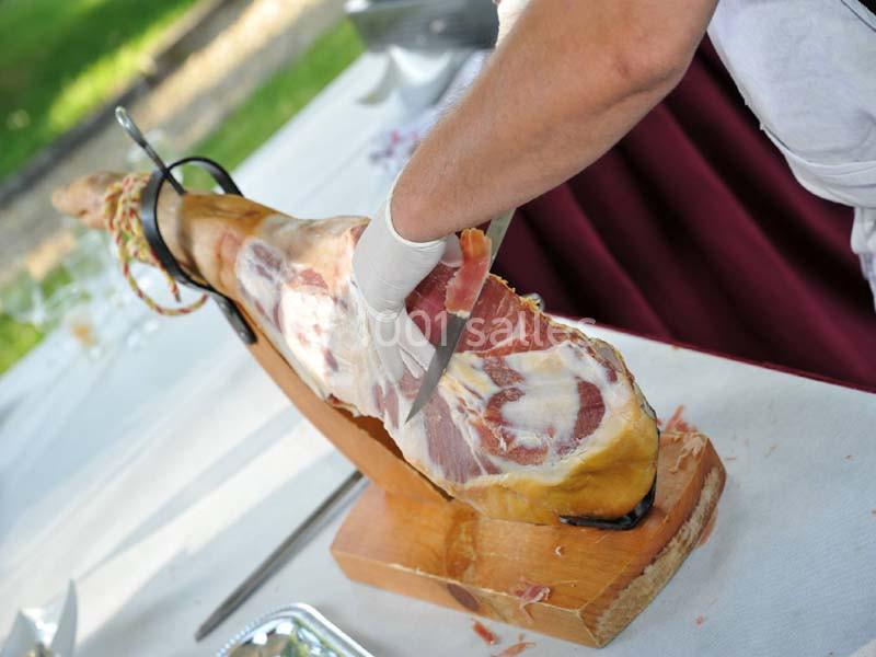 Une personne découpe finement un jambon entier posé sur un support en bois, en extérieur.