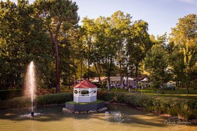 Un kiosque blanc au bord d'un étang avec une fontaine, entouré d'arbres et d'une réception en arrière-plan.