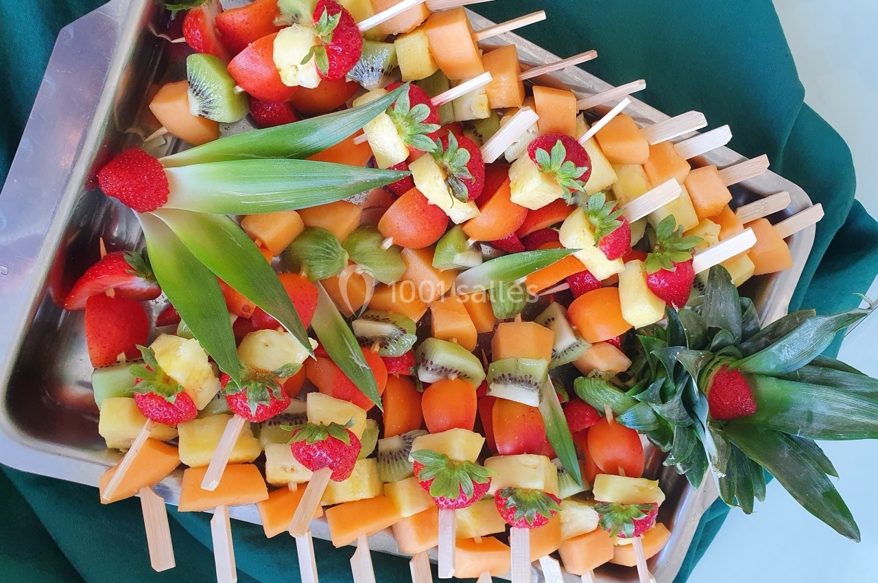 Plateau de brochettes de fruits colorés avec fraises, ananas, kiwi et melon, présenté sur un tissu vert.