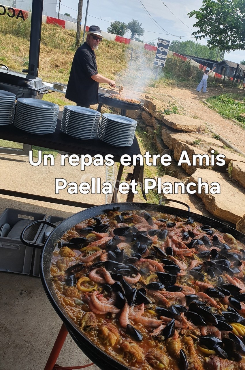 Un cuisinier prépare un repas en plein air avec une grande paella garnie de fruits de mer au premier plan.