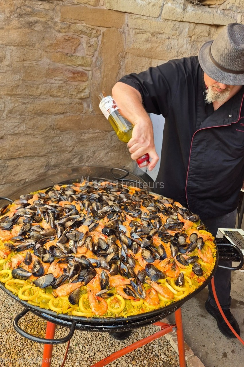 Un chef prépare une grande paella garnie de moules, crevettes et calamars, en extérieur devant un mur en pierre.