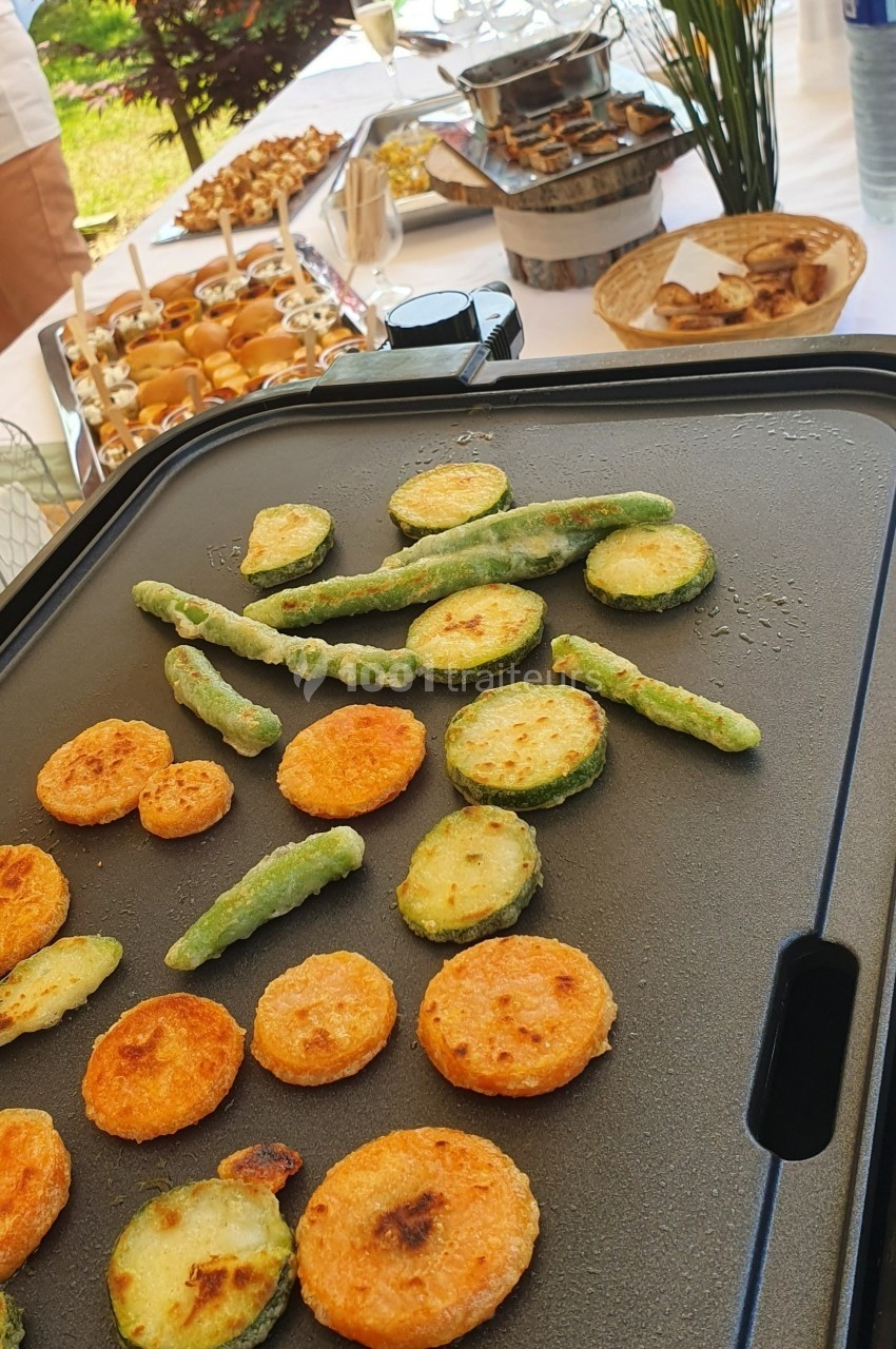 Légumes frits en train de cuire sur une plancha, avec un buffet varié en arrière-plan sur une table en extérieur.