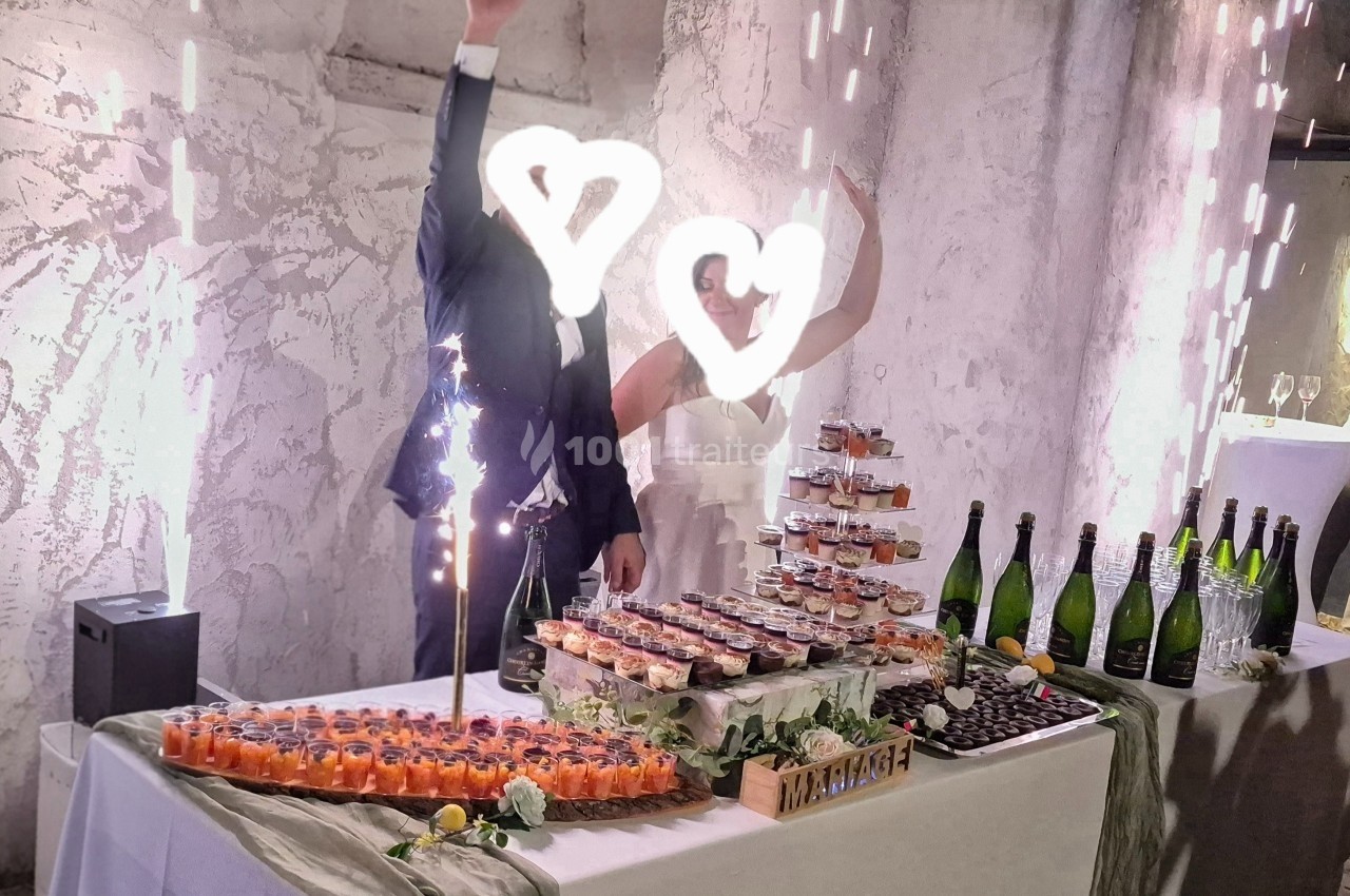 Un couple en tenue de mariage salue devant une table garnie de desserts et de bouteilles, décorée pour une réception.