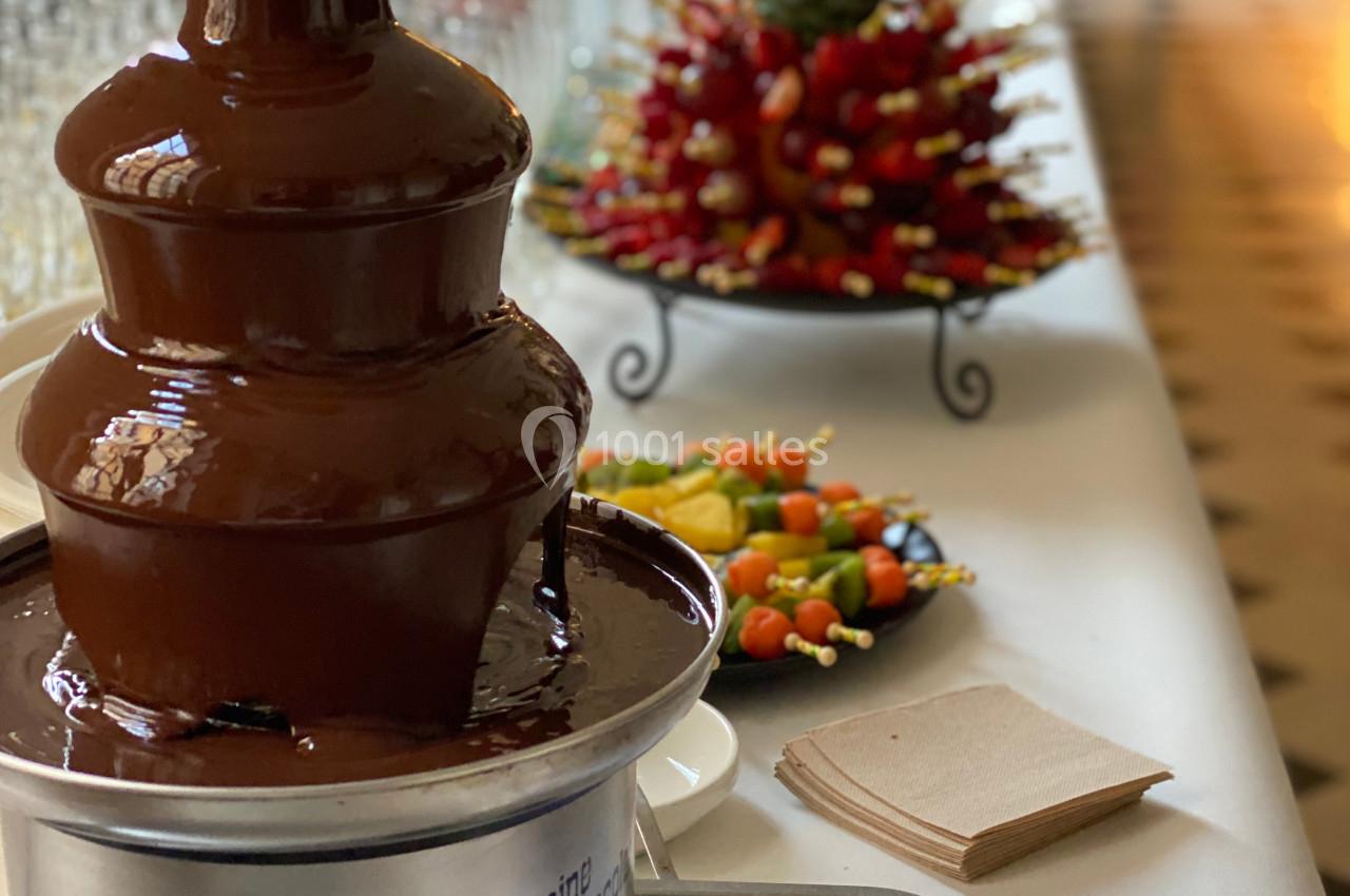 Fontaine à chocolat sur une table de buffet avec des fruits frais et des brochettes en arrière-plan.