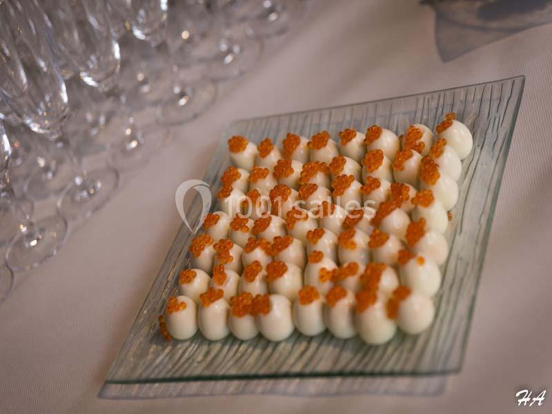 Assiette de bouchées blanches garnies de perles orange, présentée sur un plat en verre avec des verres en arrière-plan.