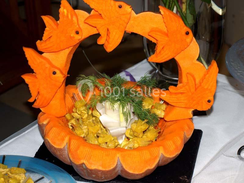 Sculpture comestible en légumes représentant des poissons orange entourant un plat garni de brochettes et d'herbes.