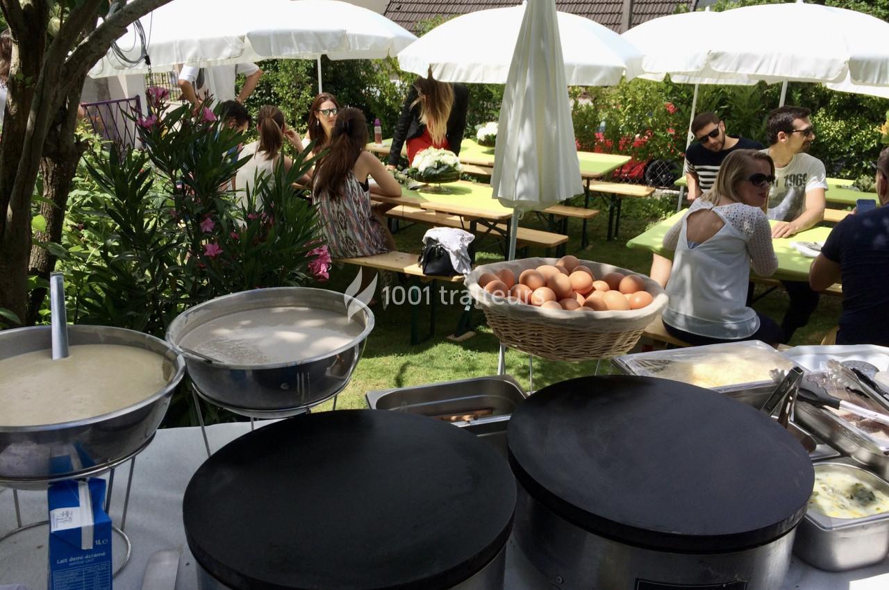 Stand de crêpes en extérieur avec des tables de convives sous des parasols dans un jardin verdoyant.