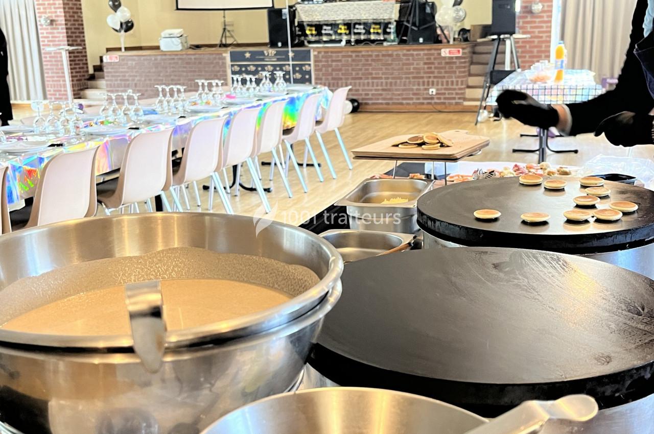 Préparation de mini-crêpes sur une crêpière dans une salle aménagée avec des tables dressées pour un événement.