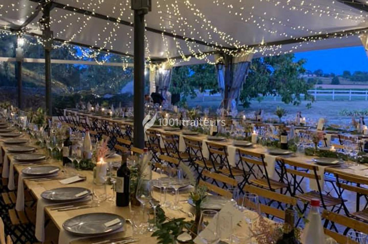 Tables décorées pour un dîner en plein air sous une tente illuminée par des guirlandes lumineuses, avec vue sur la campagne.