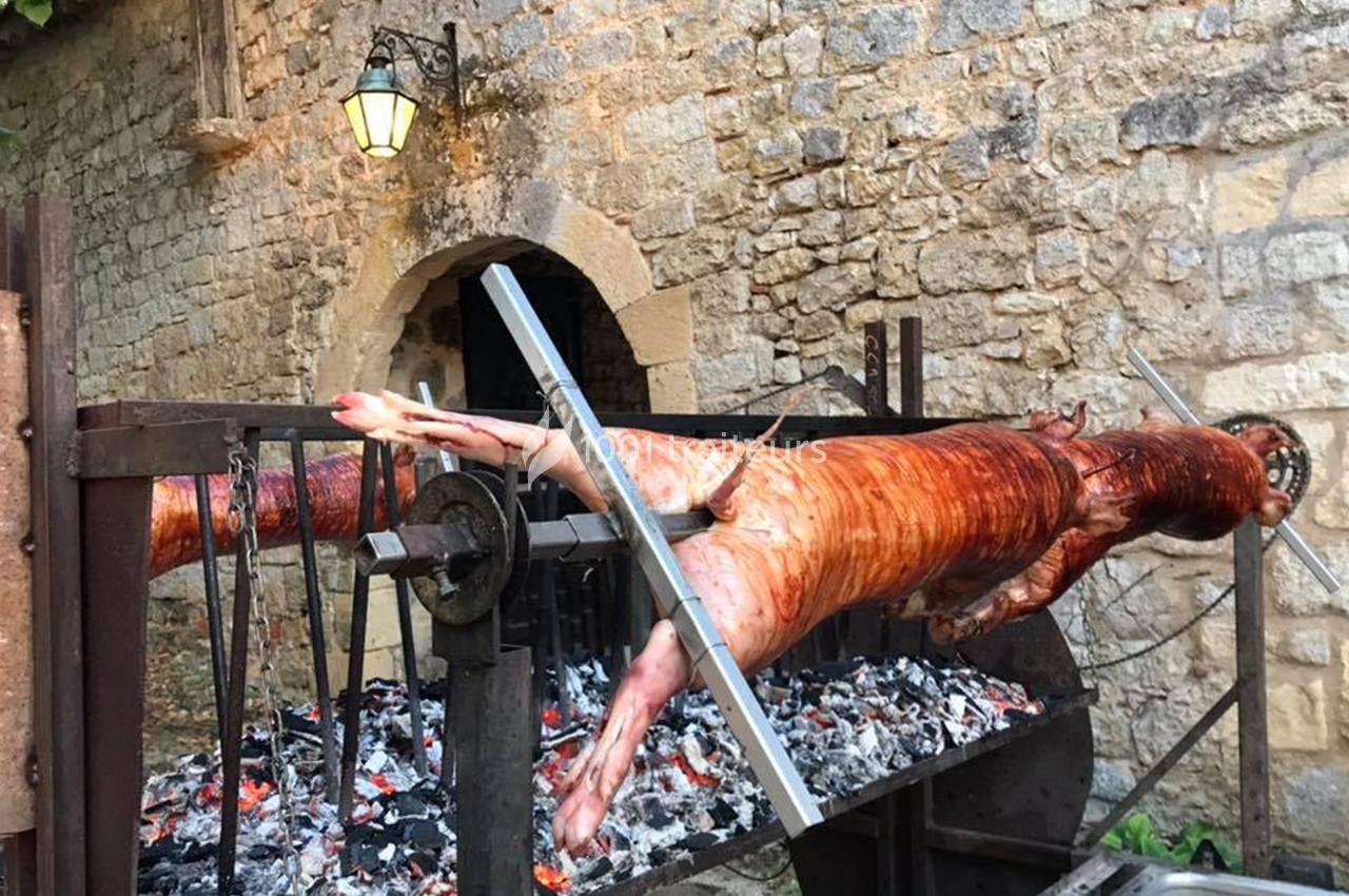 Un cochon entier rôti à la broche au-dessus de braises, devant un mur en pierre et une porte voûtée.
