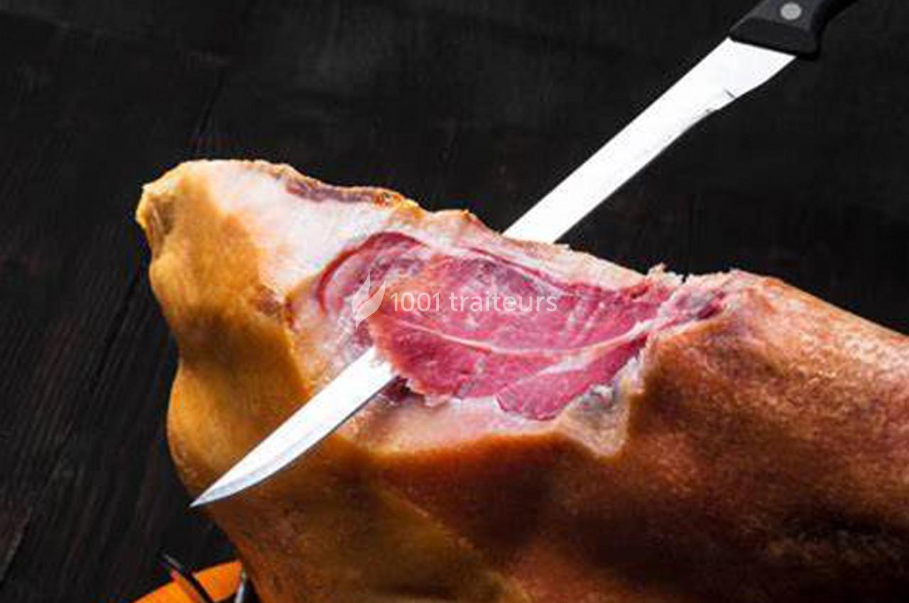 Un couteau tranche une pièce de jambon cru sur un fond sombre.