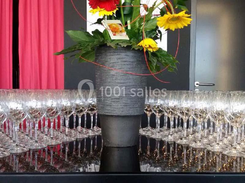 Un vase gris avec des fleurs colorées posé sur une table entourée de nombreux verres à pied.