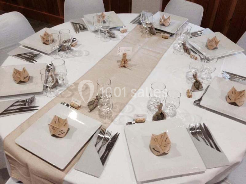 Table dressée avec nappes blanches, chemins de table beige, assiettes carrées, verres et pliages en papier décoratifs.