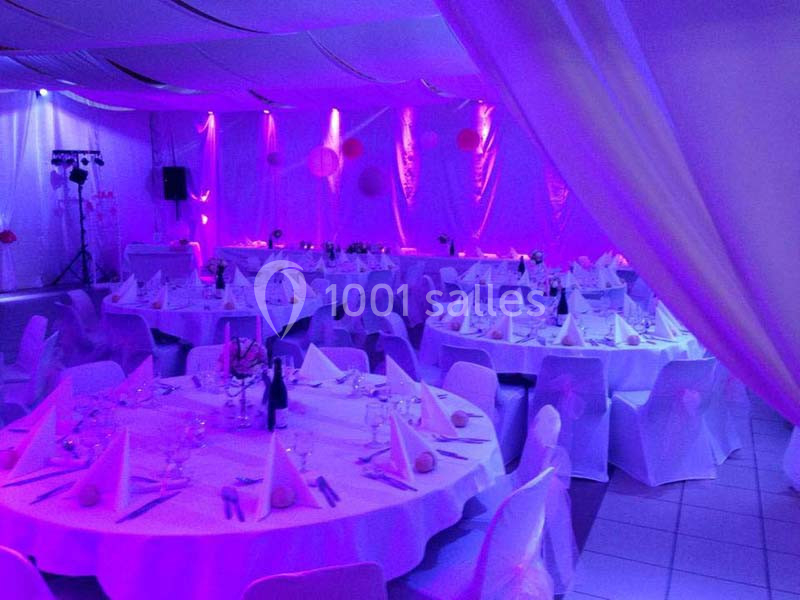 Salle de réception décorée avec des nappes blanches, des tables rondes dressées et un éclairage violet tamisé.