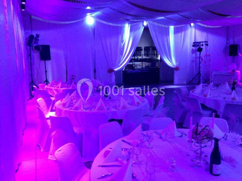 Salle de réception décorée avec des tables rondes dressées, éclairage violet et scène avec matériel audio.