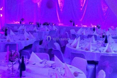 Salle de réception décorée avec des nappes blanches, des tables rondes dressées et un éclairage violet.