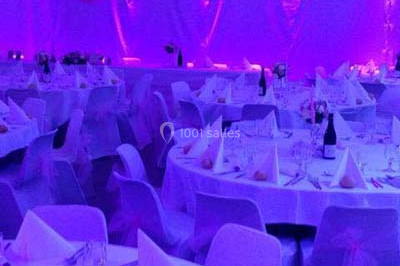 Salle de réception décorée avec des nappes blanches, des tables rondes dressées et un éclairage violet.