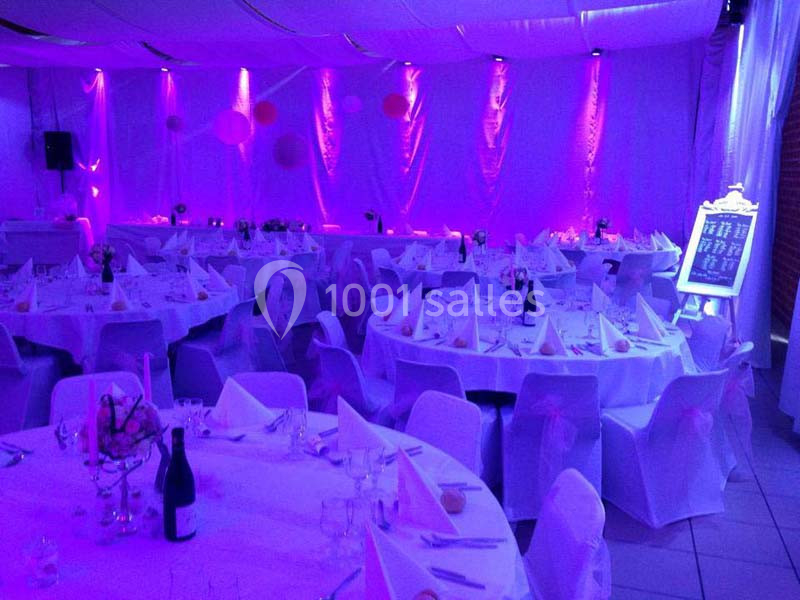 Salle de réception décorée avec des nappes blanches, des tables rondes dressées et un éclairage violet.