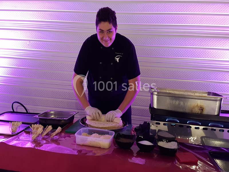 Un cuisinier prépare des plats sur une table avec des ingrédients et des ustensiles, devant un fond éclairé en violet.