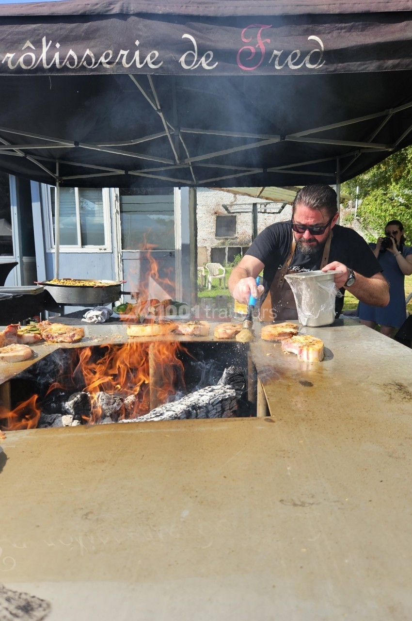 Un cuisinier prépare des plats sur un grill en plein air avec des flammes sous un auvent noir.