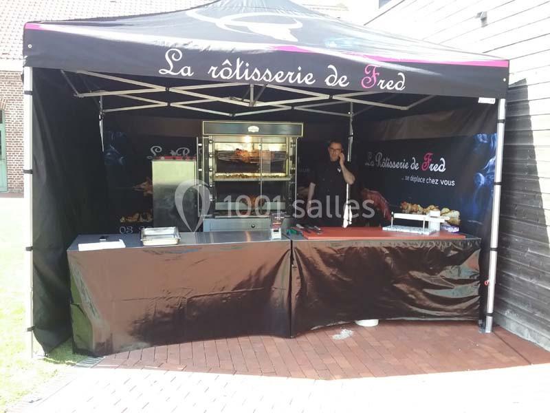 Stand de rôtisserie avec un four à poulets grillés, un comptoir et un vendeur sous une tente noire.