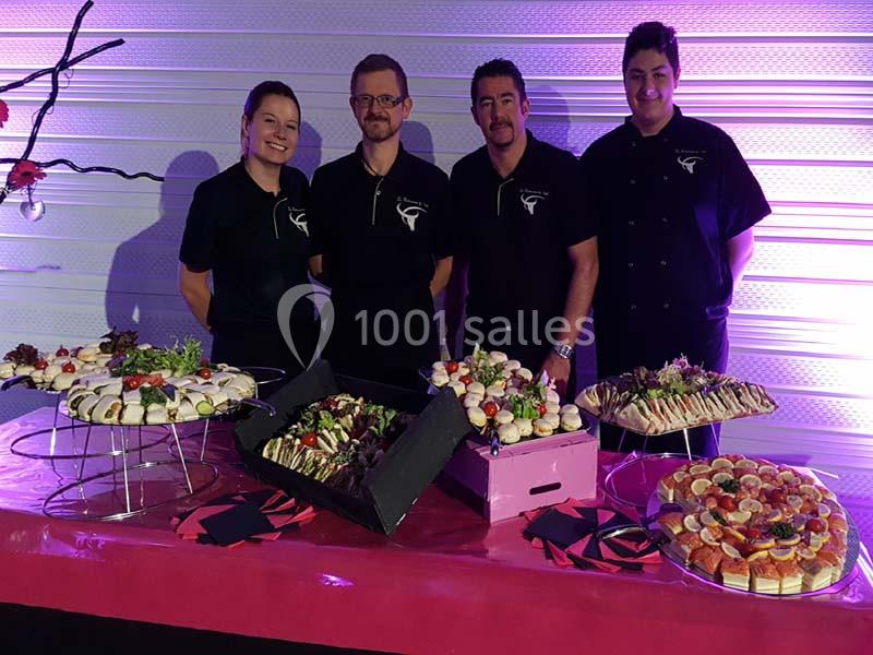 Quatre personnes posent derrière une table garnie de plateaux de bouchées et de plats variés dans un cadre éclairé en violet.