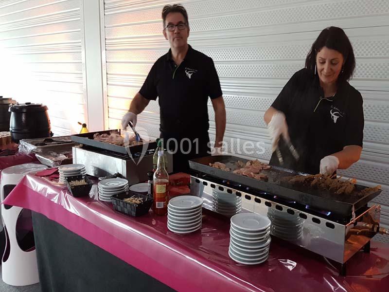 Deux personnes préparent des plats sur des planchas lors d'un événement, avec des assiettes et condiments disposés devant.