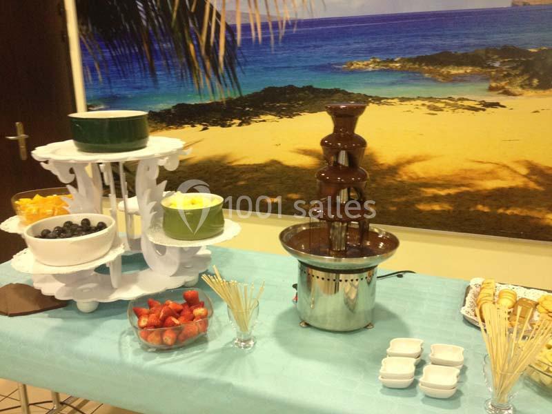 Fontaine de chocolat entourée de fruits frais, biscuits et brochettes en bois sur une table décorée, avec un fond de plage.