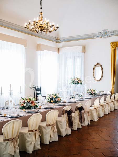Salle élégante avec une longue table dressée, décorée de fleurs et de chaises couvertes, éclairée par un lustre.