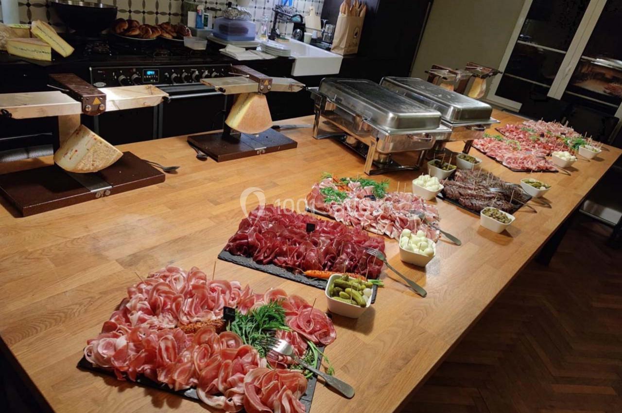 Table en bois garnie de charcuteries variées, fromages, cornichons et plats chauds dans un cadre de cuisine.