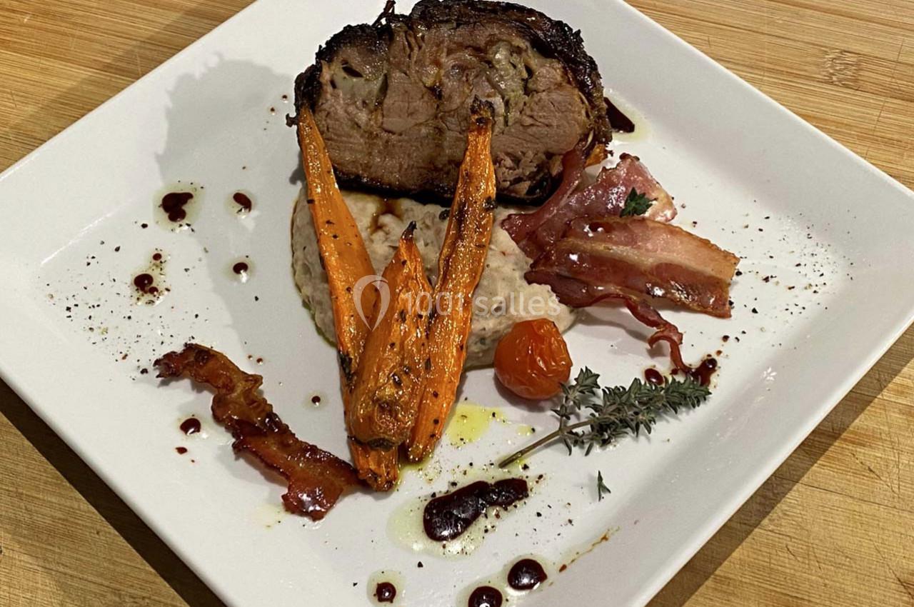 Assiette garnie d'un rôti de viande, purée, carottes rôties, bacon, tomate cerise et herbes sur une table en bois.