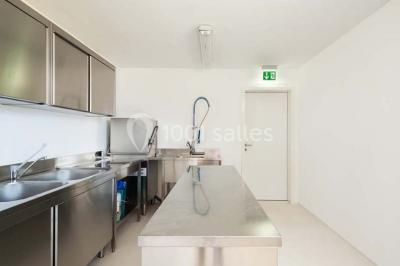 Cuisine professionnelle avec plans de travail en inox, évier, robinet flexible et porte de sortie au fond.