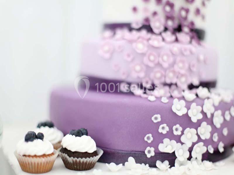 Gâteau à étages violet décoré de fleurs blanches en sucre, accompagné de cupcakes garnis de crème et de myrtilles.