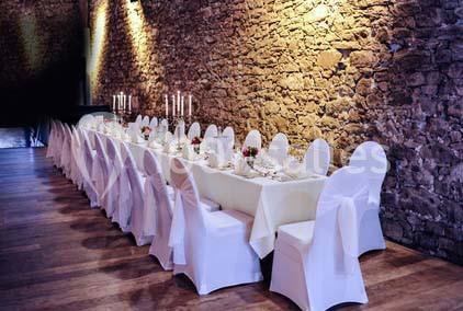 Table longue décorée pour un dîner, avec nappes blanches, chaises houssées et chandeliers, dans une salle en pierre.