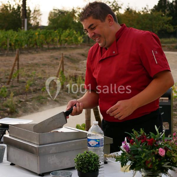 Un chef en veste rouge cuisine en extérieur près de vignes, utilisant une plaque chauffante.