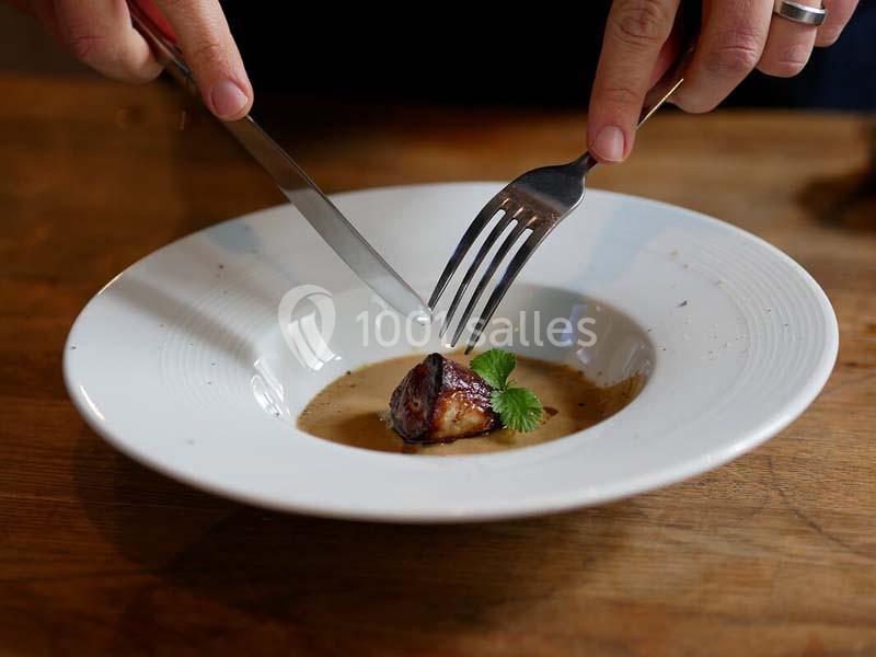 Une personne coupe un petit morceau de viande dans une assiette blanche avec une sauce et une garniture verte.