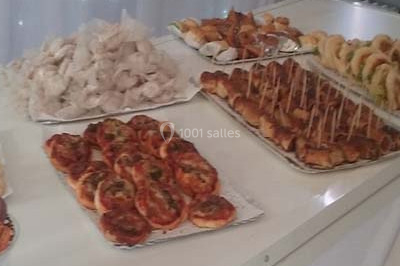 Un homme en tenue traditionnelle sert du thé près d'une table garnie de pâtisseries et de verres dans une salle lumineuse.
