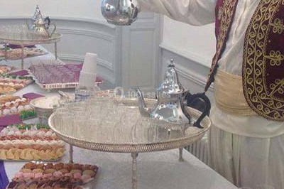 Un homme en tenue traditionnelle sert du thé près d'une table garnie de pâtisseries et de verres dans une salle lumineuse.