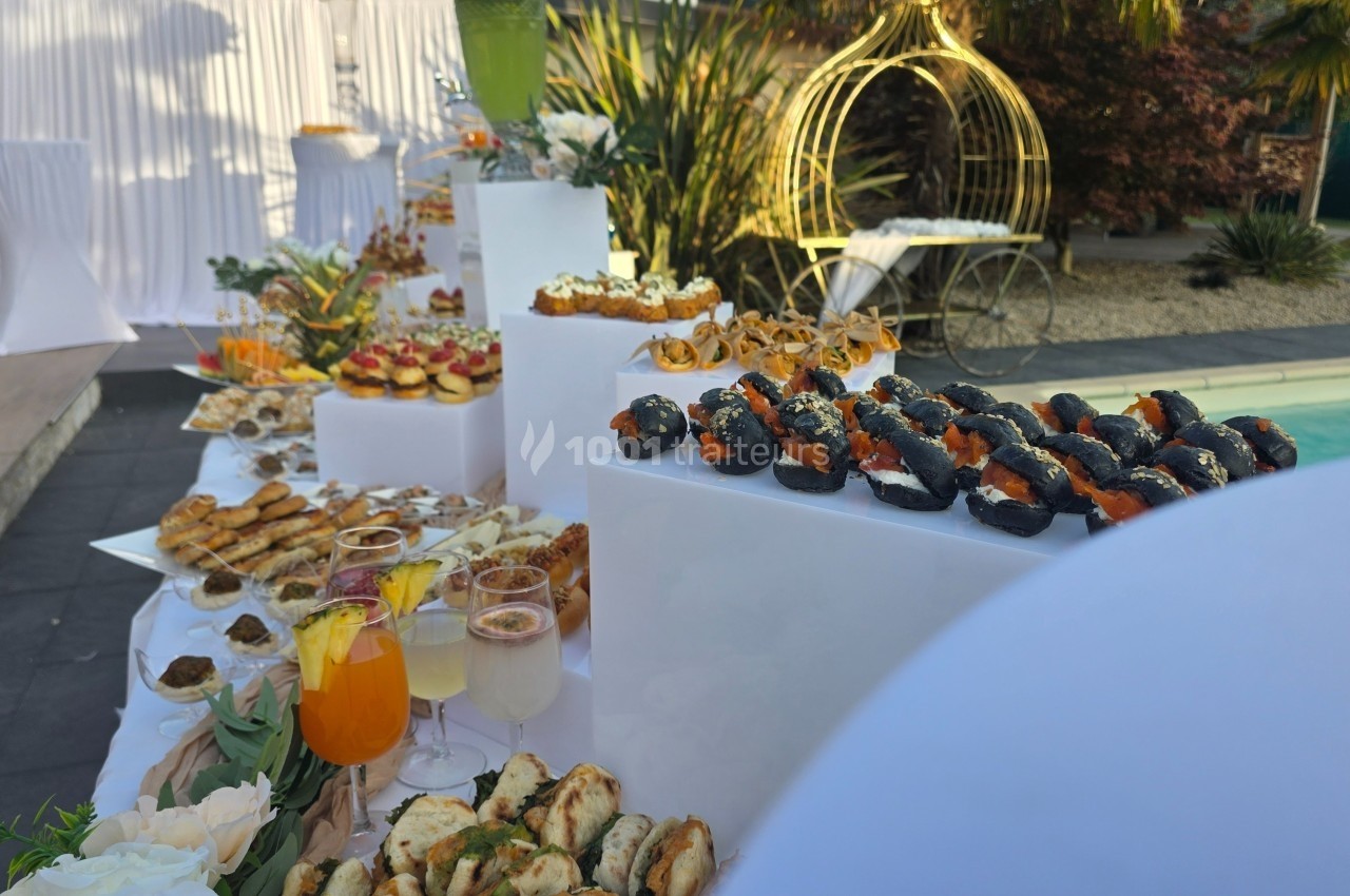 Buffet en extérieur avec divers amuse-bouches, cocktails et décorations élégantes près d'une piscine.