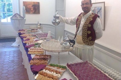 Un homme en tenue traditionnelle sert du thé près d'une table garnie de pâtisseries et de verres dans une salle lumineuse.