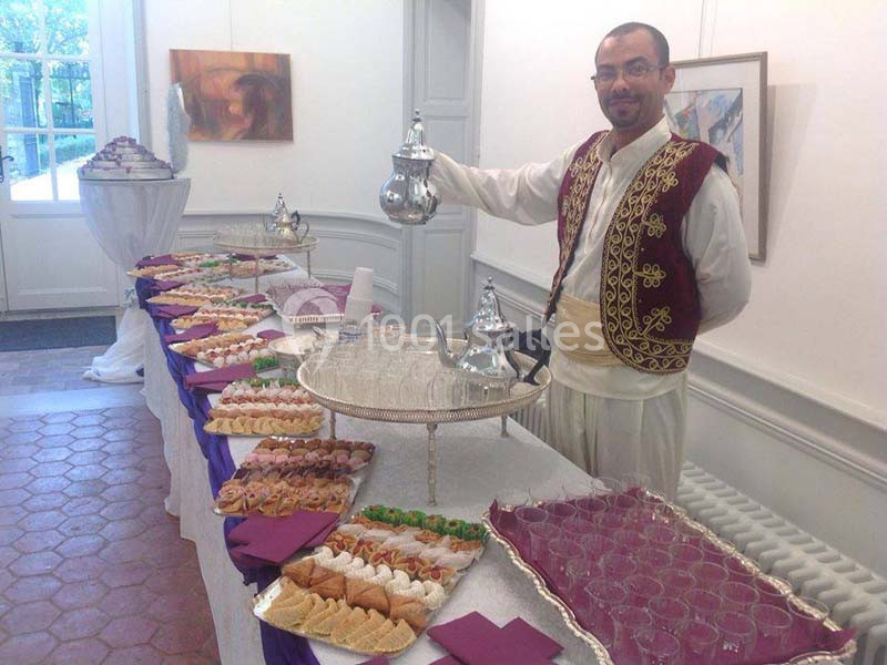 Un homme en tenue traditionnelle sert du thé à côté d'une table garnie de pâtisseries et de verres dans une salle lumineuse.