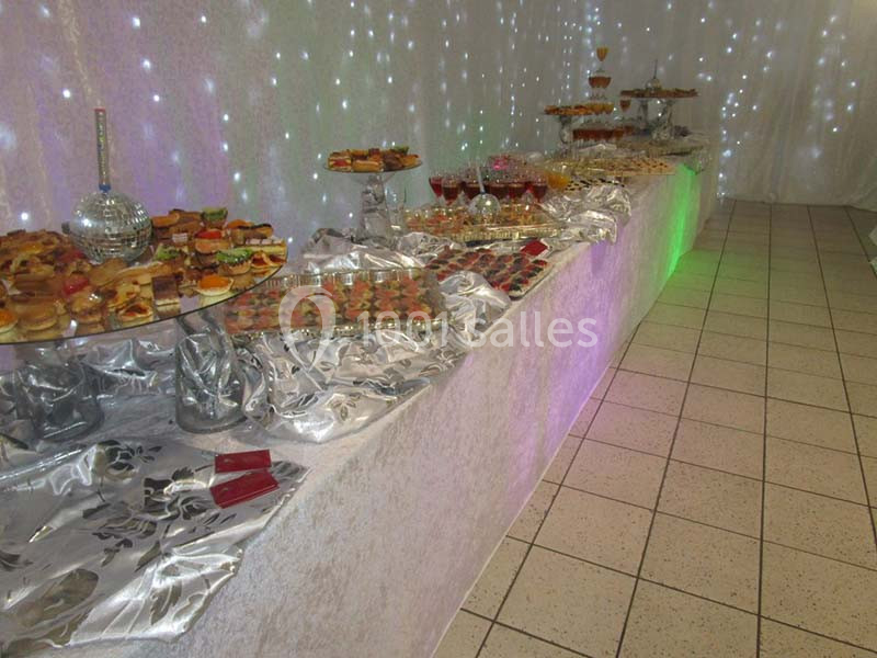 Buffet varié avec amuse-bouches, desserts et décorations lumineuses sur une longue table recouverte de nappes argentées.