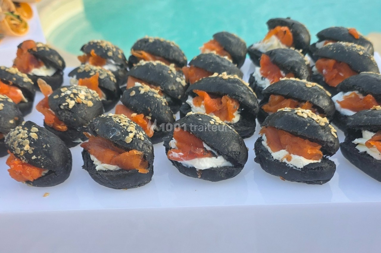 Petits pains noirs garnis de fromage frais et de saumon fumé, présentés sur un plateau près d'une piscine.