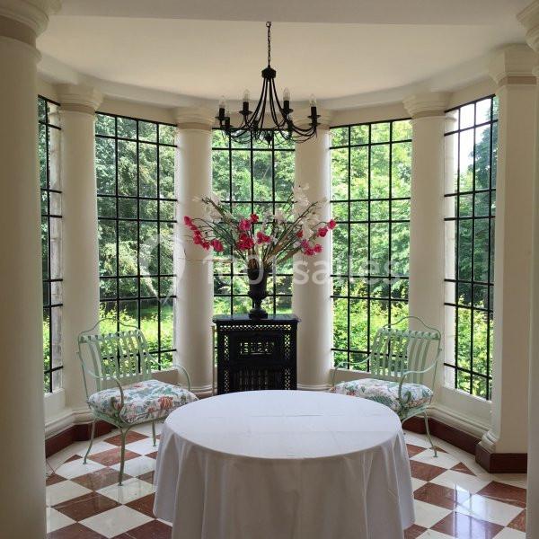Salle lumineuse avec baies vitrées, colonnes, table ronde blanche, deux chaises et un vase de fleurs colorées.