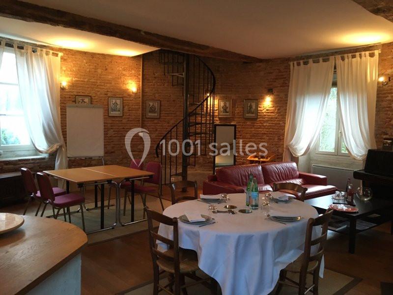 Salle chaleureuse avec murs en briques, table dressée, canapé rouge, escalier en colimaçon et grandes fenêtres.