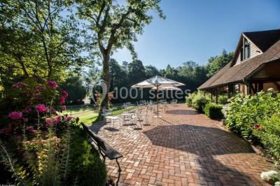 Table et chaises en bois disposées sur du gravier, face à un jardin verdoyant avec buissons et pelouse.