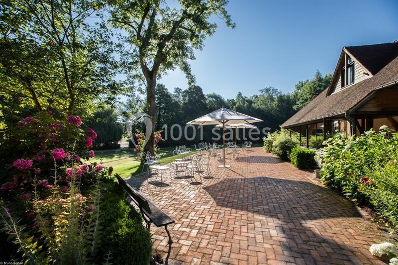 Terrasse en pavés avec tables, chaises et parasols, entourée de verdure et fleurs sous un ciel dégagé.